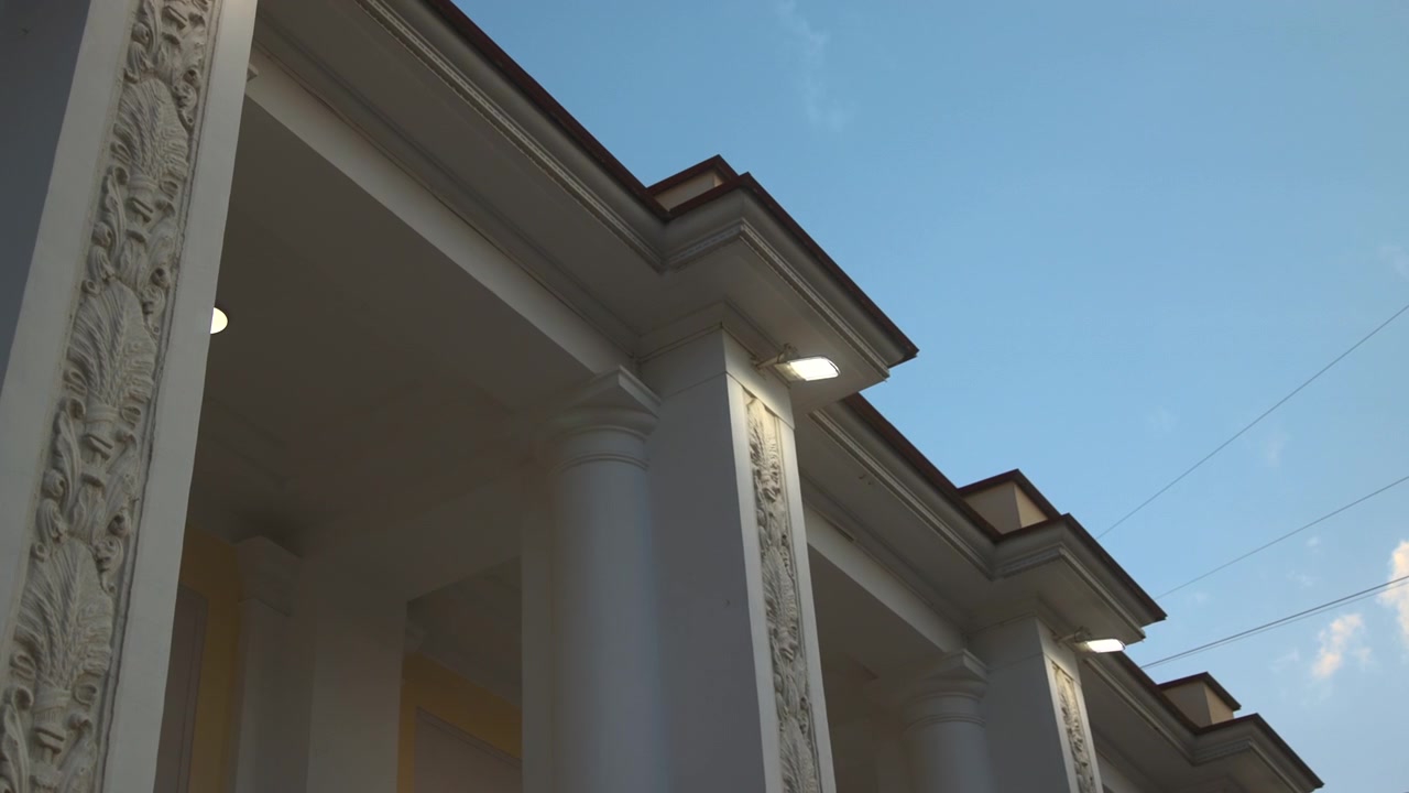Upward view of historic institutional facade showing tall rounded columns, carved vertical relief, layered roofline, corner detail and soft exterior lighting against deep blue dusk sky
