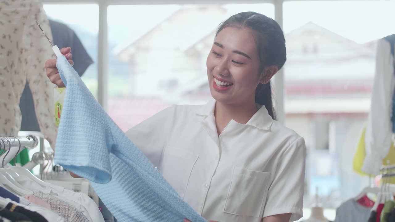 젊은 아시아 여성이 파란색 옷을 선택하고 부티크에서 쇼핑하는 동안 카메라에 미소 짓고 있습니다.