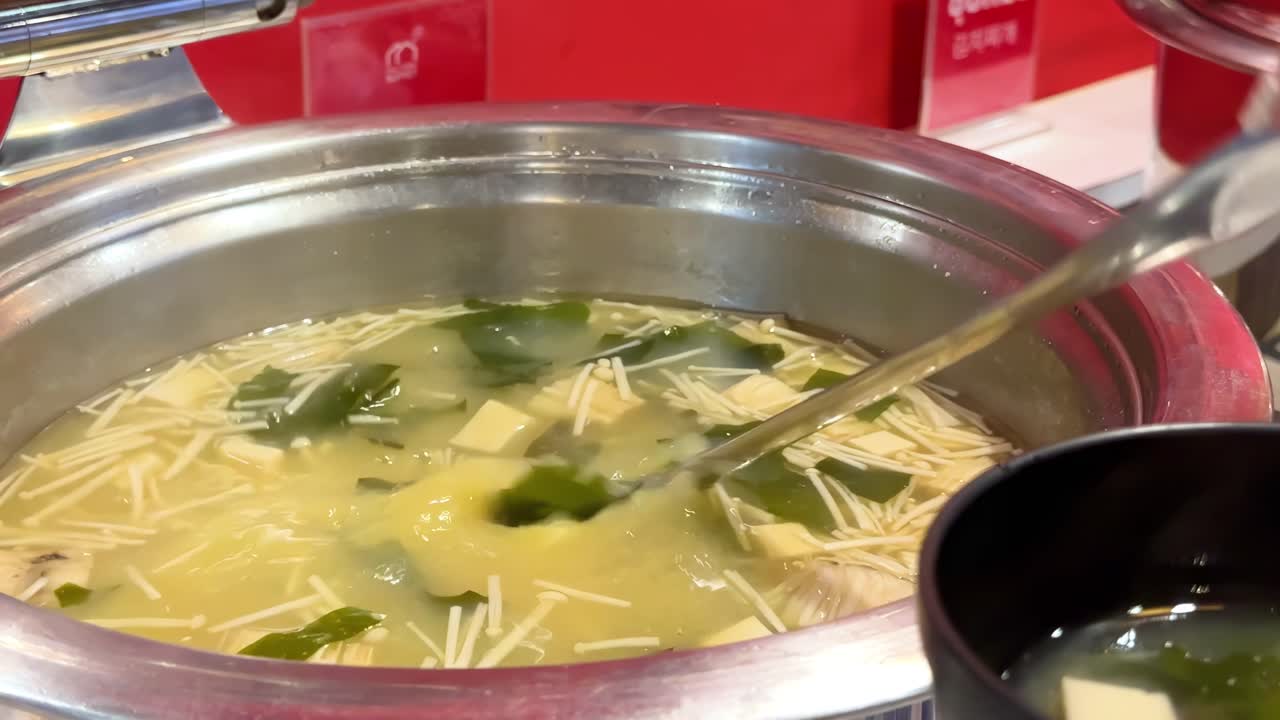 A ladle is used to serve miso soup with tofu and seaweed into a black bowl.