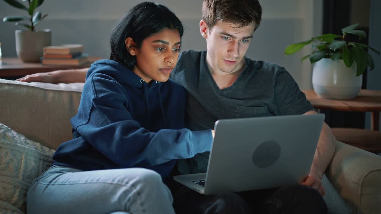 Leaning woman guiding partner, tapping laptop on sofa at home to review content, hoodie