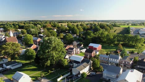 high and wide aerial overhead shot of small american town, village, establishing shot shows homes and housing along street, green trees and farmland in distance during august, north america, usa