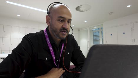 hombre calvo asiático indio en la oficina usando portátil usando auriculares, entorno de trabajo enfocado, interior, durante el día