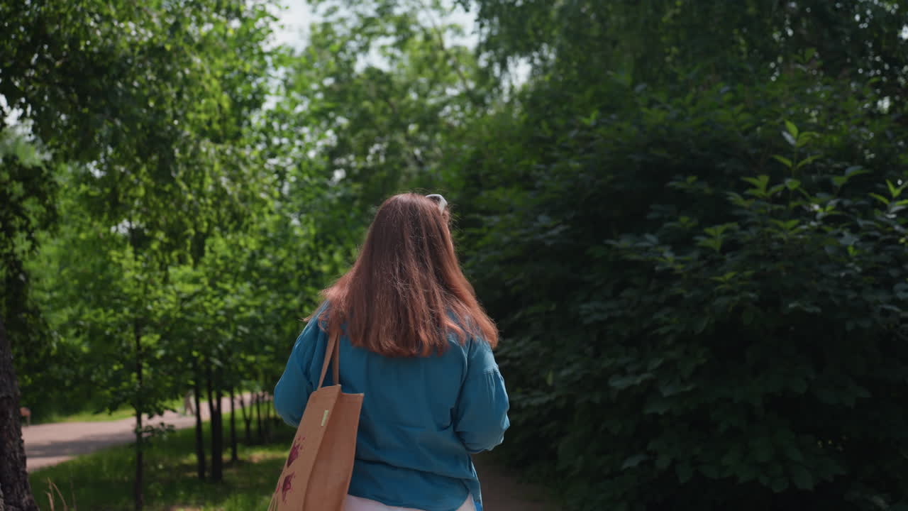 Back view of lady wearing blue top and carrying handbag walking along sunny park alley surrounded by lush green trees and soft summer light, enjoying peaceful outdoor stroll