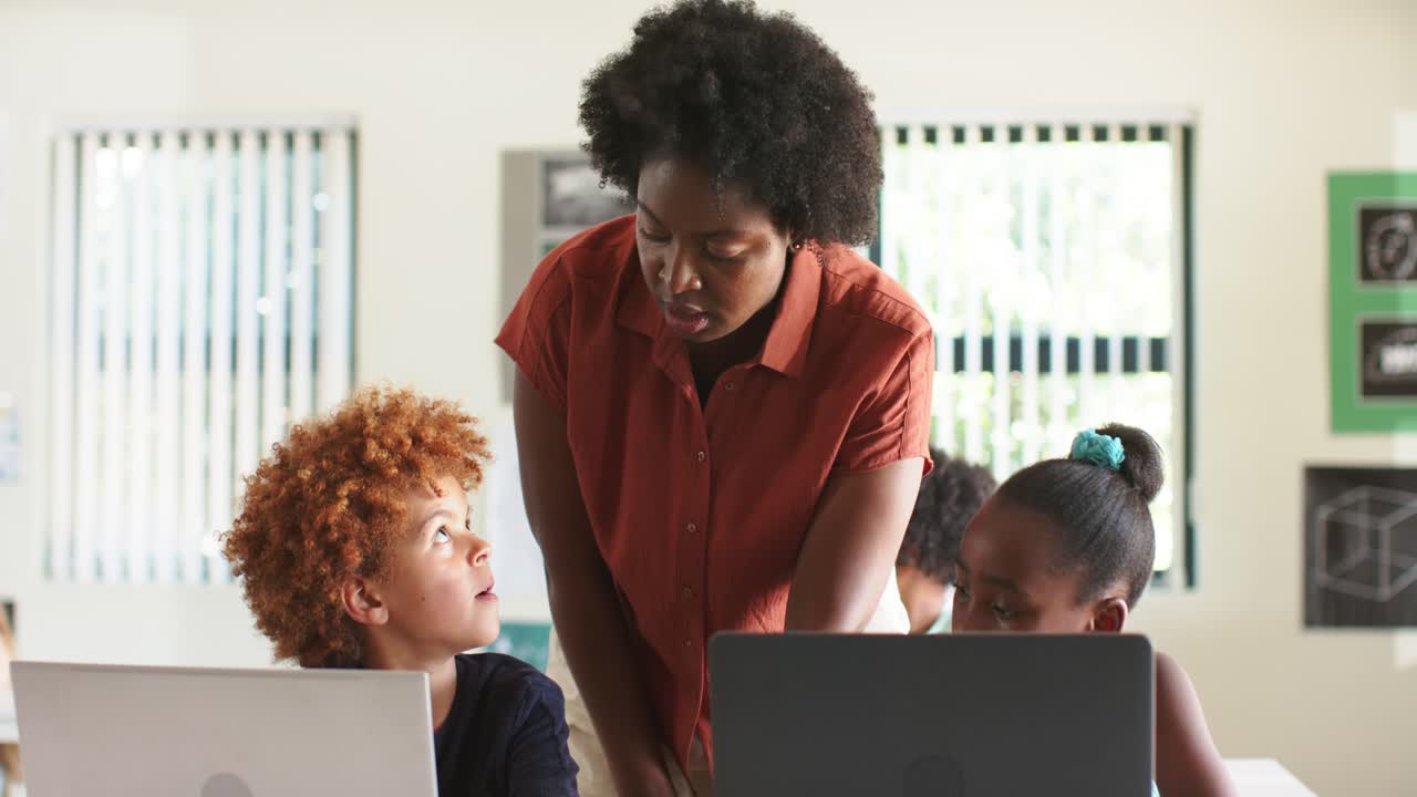 African American teacher noticing two laptops, leaning, guiding boy's keyboard in classroom to help