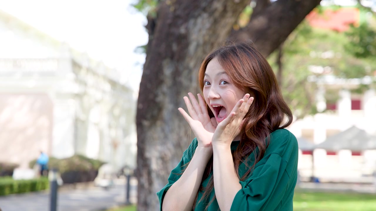 明るい日中の光の中で、驚いたアジア人女性が口を開け、顔に手を当てて反応している。