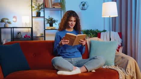 woman reading interesting book turning pages preparing for exam sitting on couch in room at home