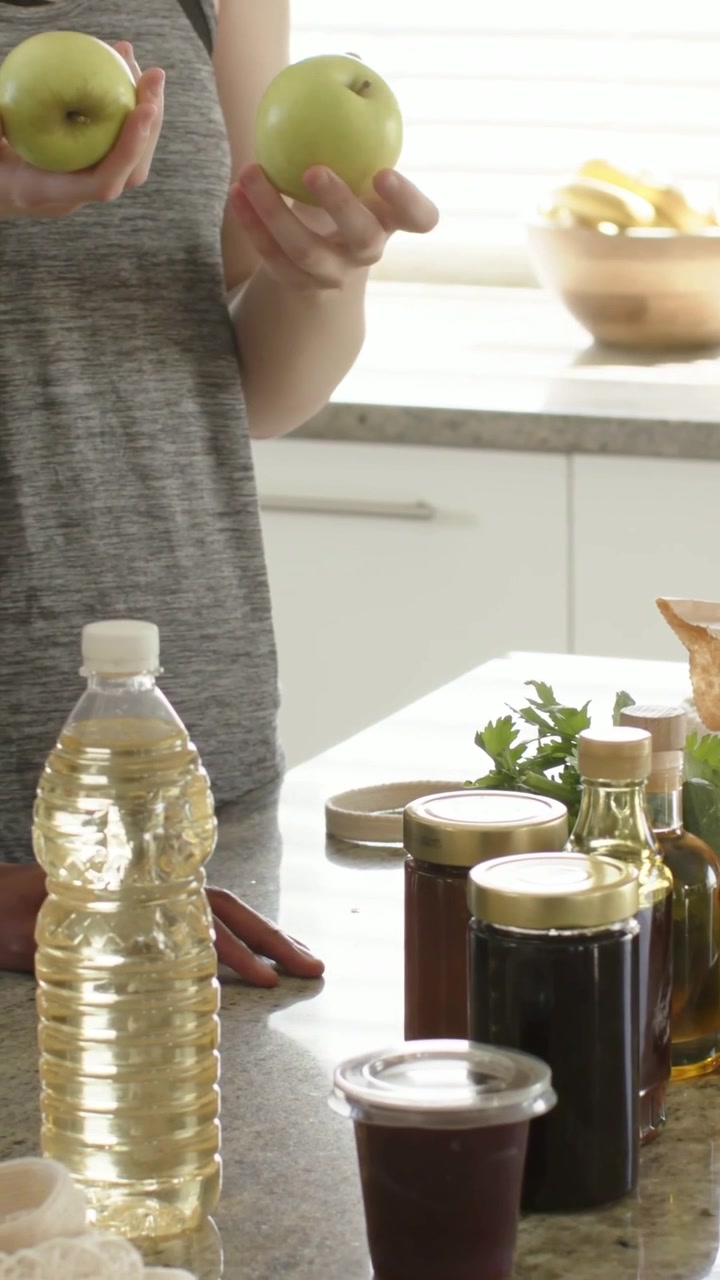 Vertical video: Holding green apples, woman in gray top comparing, arranging jars at kitchen island