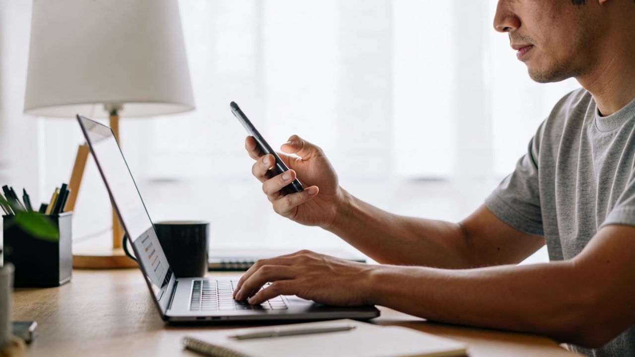Man in grijs T-shirt reageert op telefoonmelding, houdt smartphone omhoog, typt op laptop, lamp, ruimte voor tekst.