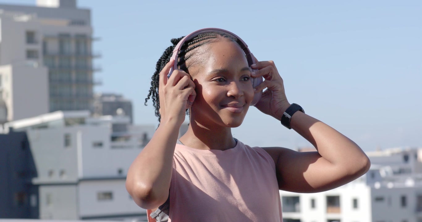Afrikansk kvinde tjekker lyserøde hovedtelefoner på et tag, justerer dem, tager dem på og starter afspilning på sit smartwatch.
