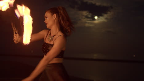 woman dancing and twirling with fire fans against a cloudy full moon lit night sky, medium shot slowmo