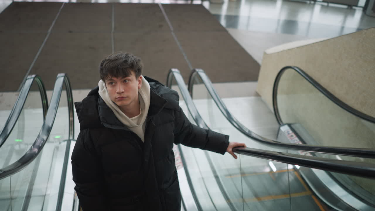 Young European boy riding escalator upward in mall wearing casual clothes holding rail with focused expression soft light casting subtle shadows blurred shoppers in background urban atmosphere smile
