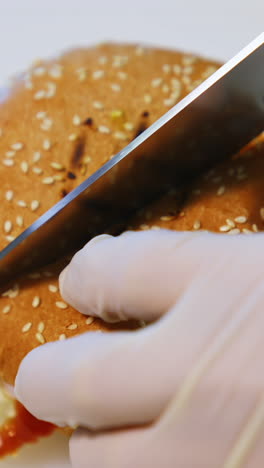 hands in gloves of a cook making burger. chef cutting ready hamburger into halves with a sharp knife on table. close-up. vertical video