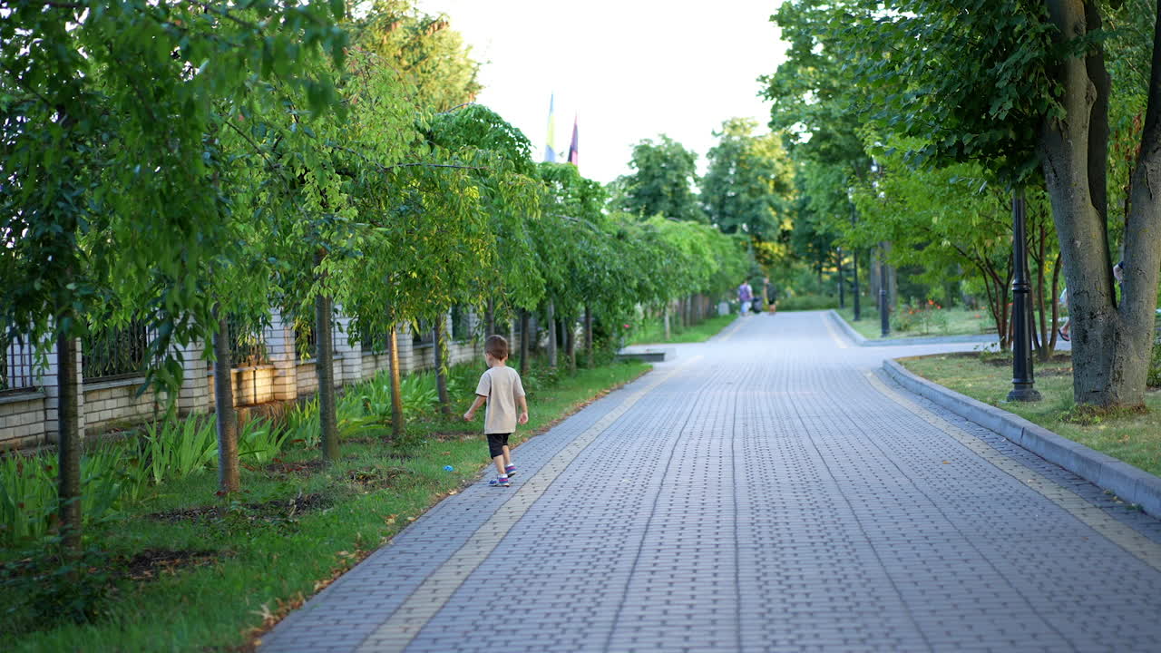 Caucasian baby boy throwing a ball on the paved alley in the park. Kid runs to catch up with the ball. Fun in summer outdoors.