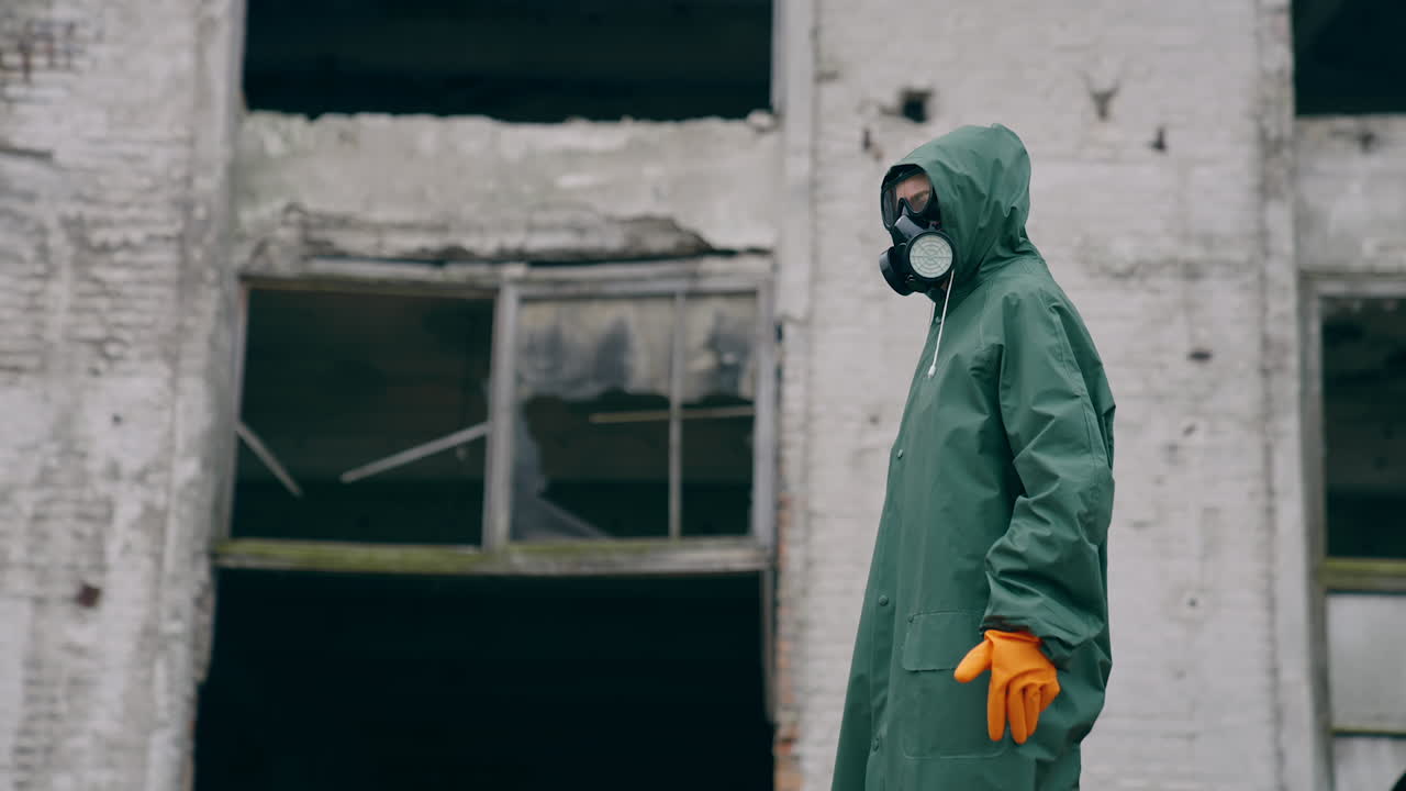 Perplexed survivor in abandoned place. Human in safety uniform and gas mask standing alone and looking to the sides on the background of destroyed buildings.