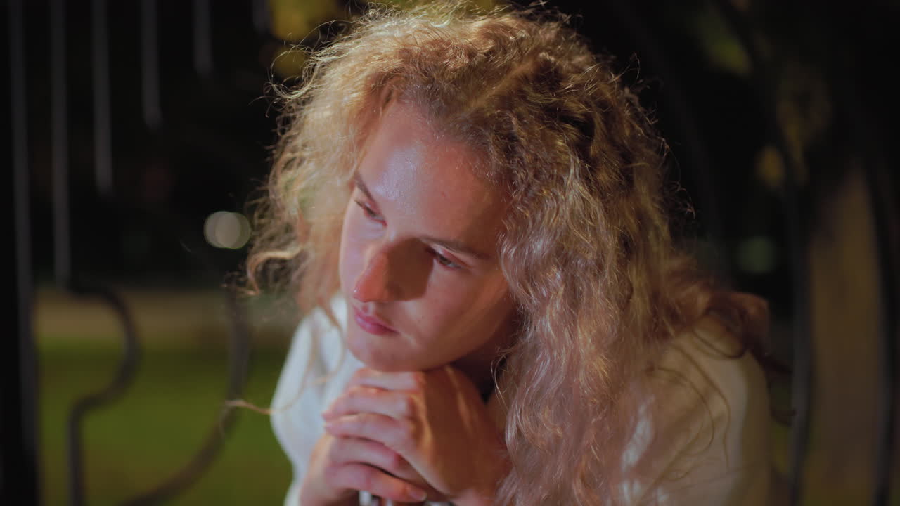 Close up of woman with curly hair in white coat feeling sleepy while seated, adjusting head on umbrella handle under warm night light glow, her face softly illuminated with tired calm expression