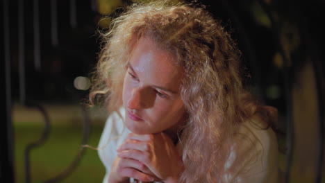 close up of woman with curly hair in white coat feeling sleepy while seated, adjusting head on umbrella handle under warm night light glow, her face softly illuminated with tired calm expression