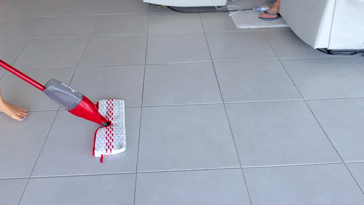 A person mops a tiled floor with a red mop in a bright, modern living room