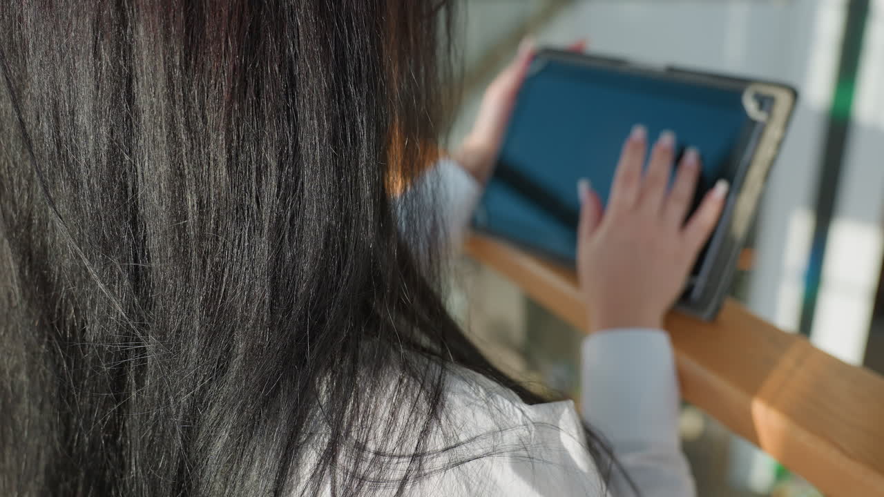 Rear view of woman with long dark hair sliding fingers across tablet screen, soft light creating subtle reflections on display, wooden rail in foreground, modern glass architecture in background