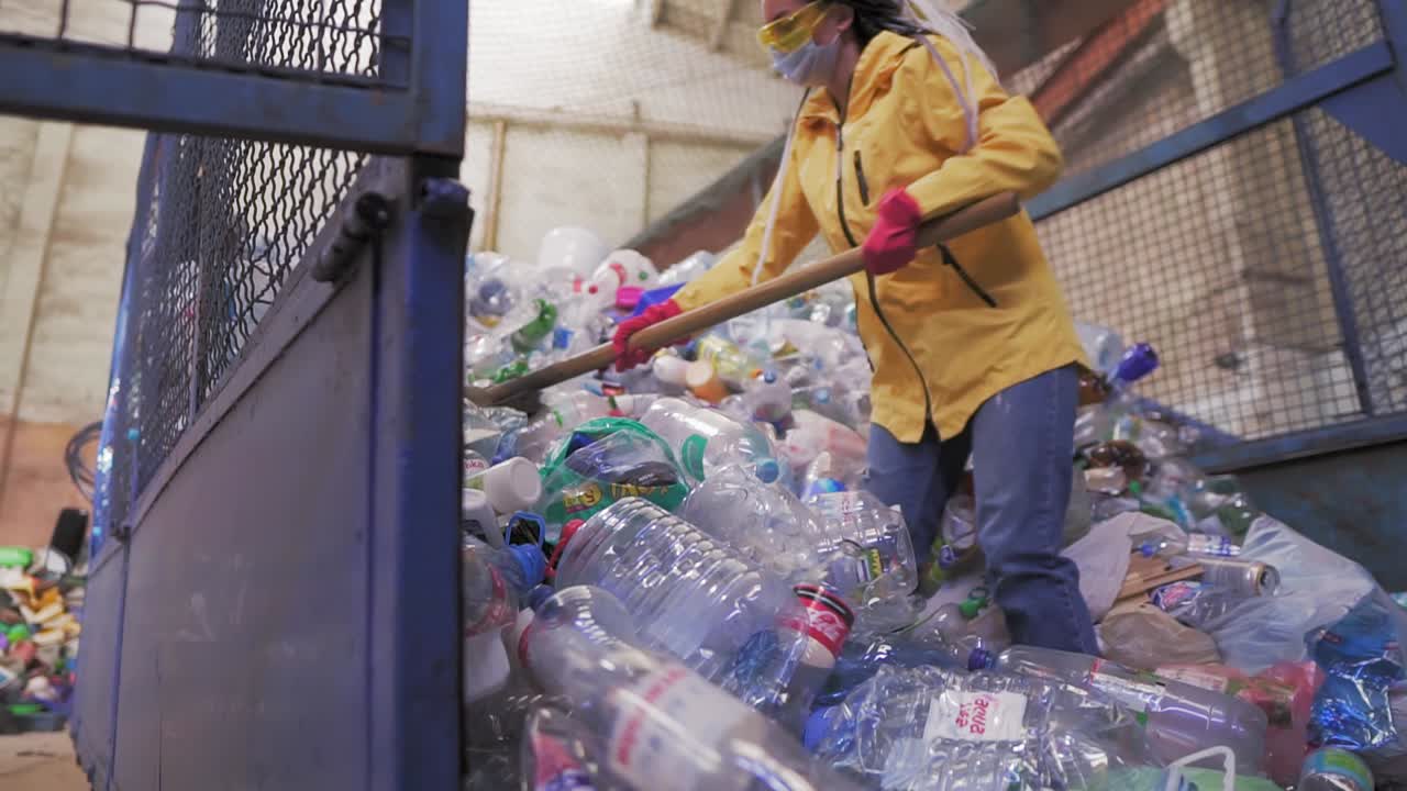Footage of a young woman in yellow jacket and gloves scoops using shovel used plastic bottles at recycling factory. Huge pile of bottles inside the car case cage. Slow motion