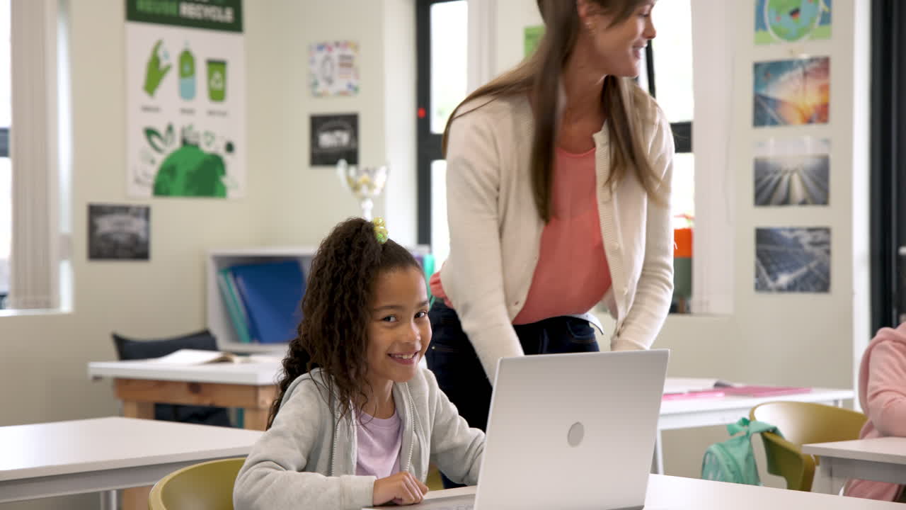 Teacher helping student with laptop in classroom, both smiling and engaged