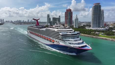 carnival cruise ship sails near south pointe park in miami beach. luxury high-rises line the waterfront, and speedboats trail behind the ship. aerial view of miami’s busy port and skyline.