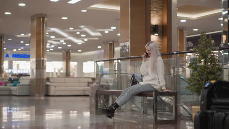 graceful mall visitor seated on bench giggling into smartphone call, playfully lifting leg while wearing knit sweater, denim and chunky black boots against backdrop of glossy tiled floor