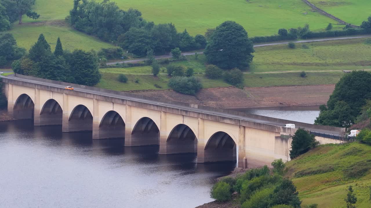 A single car drives across a large arched bridge spanning Ladybower Reservoir in Hope Valley, England, captured in a steady, wide aerial shot with soft daylight