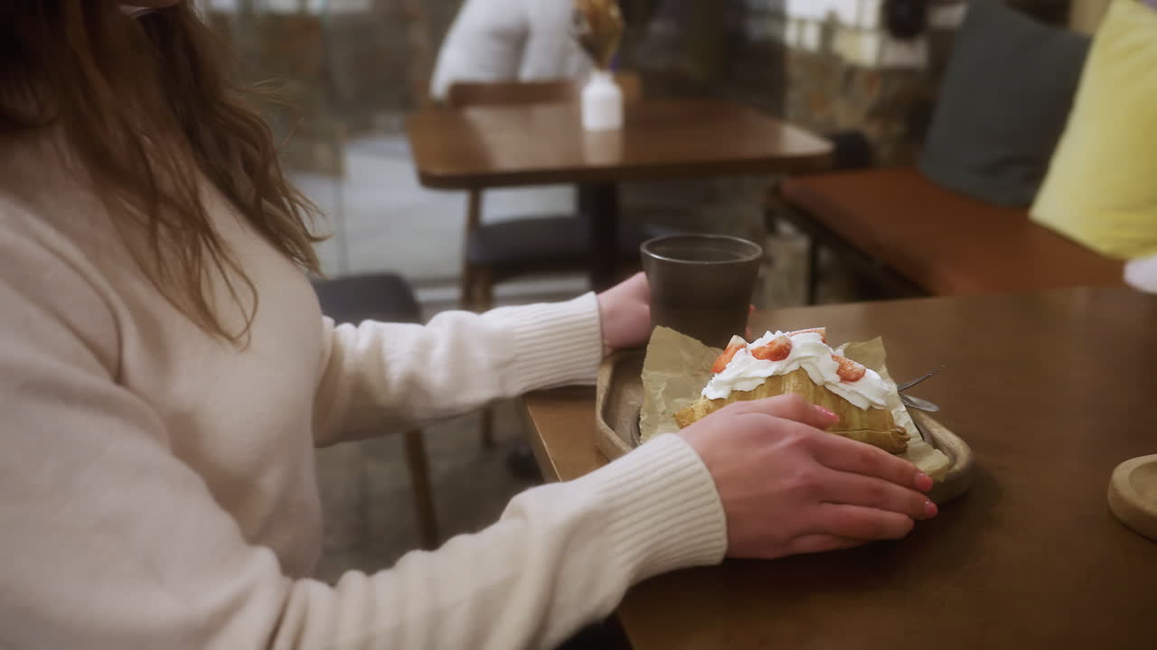 Close-up of female in beige shirt, holding coffee cup and croissant in cozy cafe, sitting near window. Soft lighting creates warm, relaxed vibe with blurred cityscape visible outside