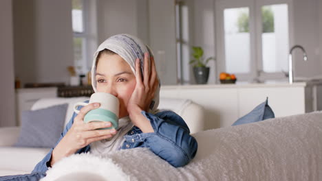 Mujer-Birracial-Feliz-Con-Hijab-Bebiendo-Té-En-El-Sofá-De-Casa-Con-Espacio-Para-Copiar,-Cámara-Lenta