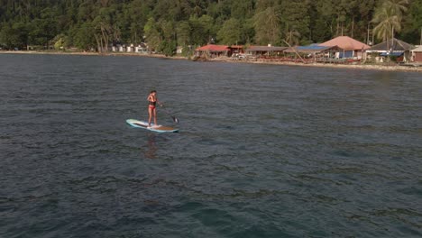 Aerial-drone-view-of-Asian-woman-with-life-vest-exercising-on-a-sup-paddle-board-in-turquoise-tropical-clear-waters,-with-beach-and-coastline-in-Thailand