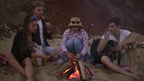 group of young and cheerful people sitting by the fire on the beach in the evening, grilling sausages and playing guitar