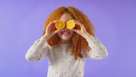 Retrato-De-Estudio-De-Una-Niña-Sosteniendo-Dos-Mitades-Naranjas-Frente-A-Los-Ojos-Contra-Un-Fondo-Púrpura