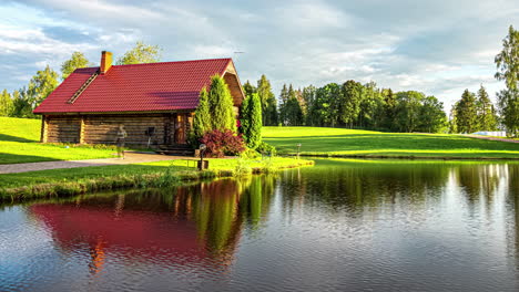 Timelapse-shot-of-a-lakeside-wooden-cottage-with-white-clouds-passing-by-over-green-grasslands-throughout-the-day