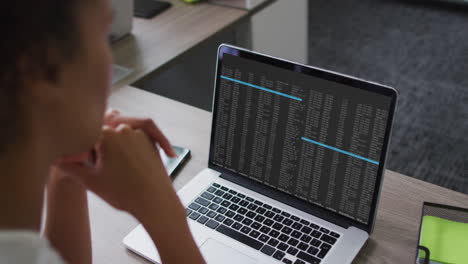 Mixed-race-woman-sitting-at-desk-watching-coding-data-processing-on-laptop-screen