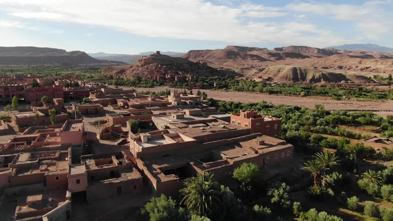 Vista aerea con drone di un tradizionale villaggio di fango circondato da un'oasi di palme e una valle verde con le Montagne dell'Atlante sullo sfondo, che mostra il paesaggio rurale e l'ambiente desertico in Marocco.