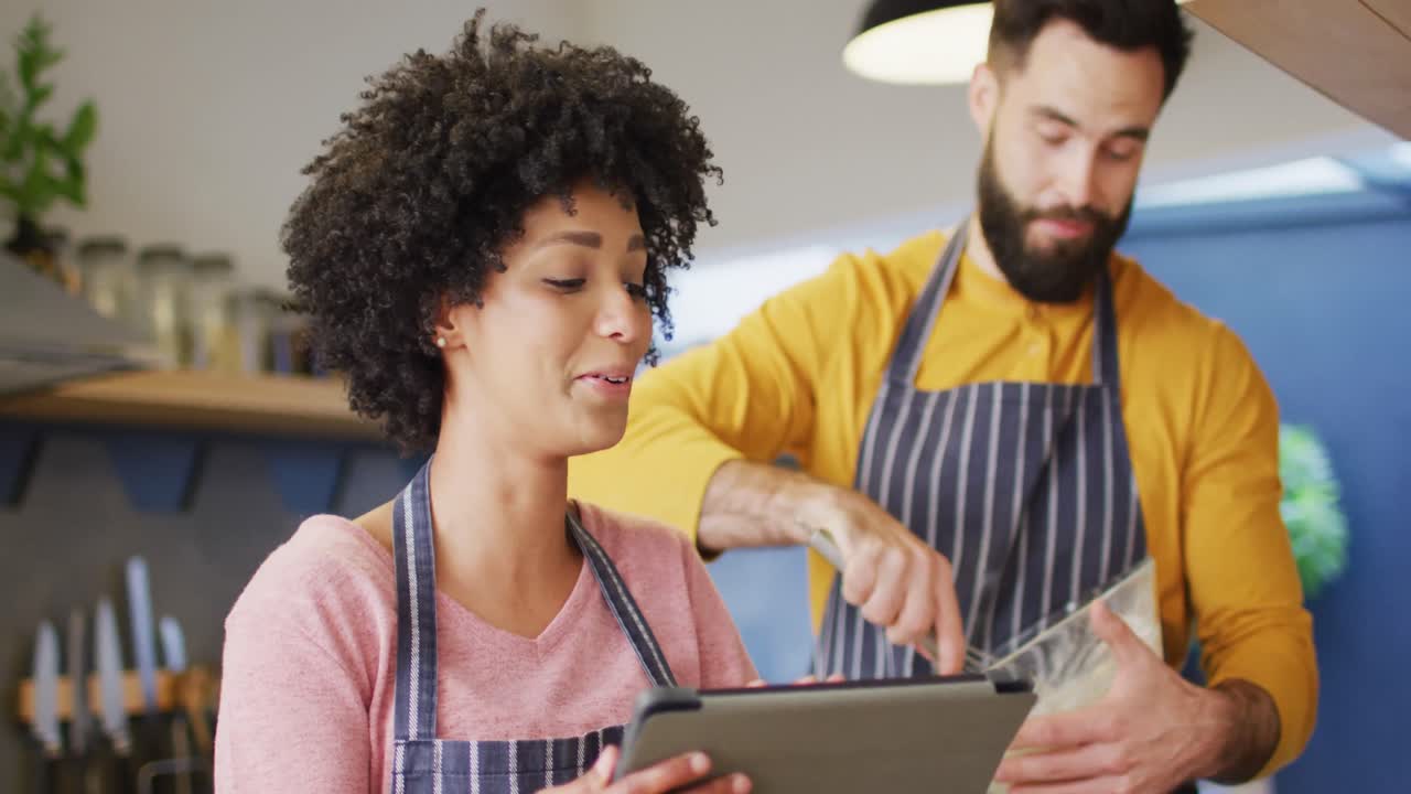 video de una feliz pareja diversa en delantales usando tableta y horneando en la cocina en casa