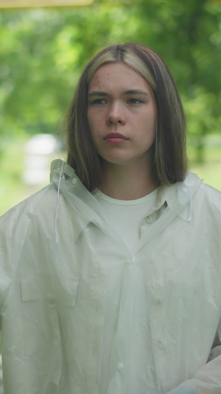 반투명한 레인코트를 입은 금발의 여인이 자전거를 타고 포장된 길을 고, 상적인 표정으로 잠시 멈추고, 배경에는 흐릿한 나무, 풀, 멀리 있는 건물들이 있습니다.