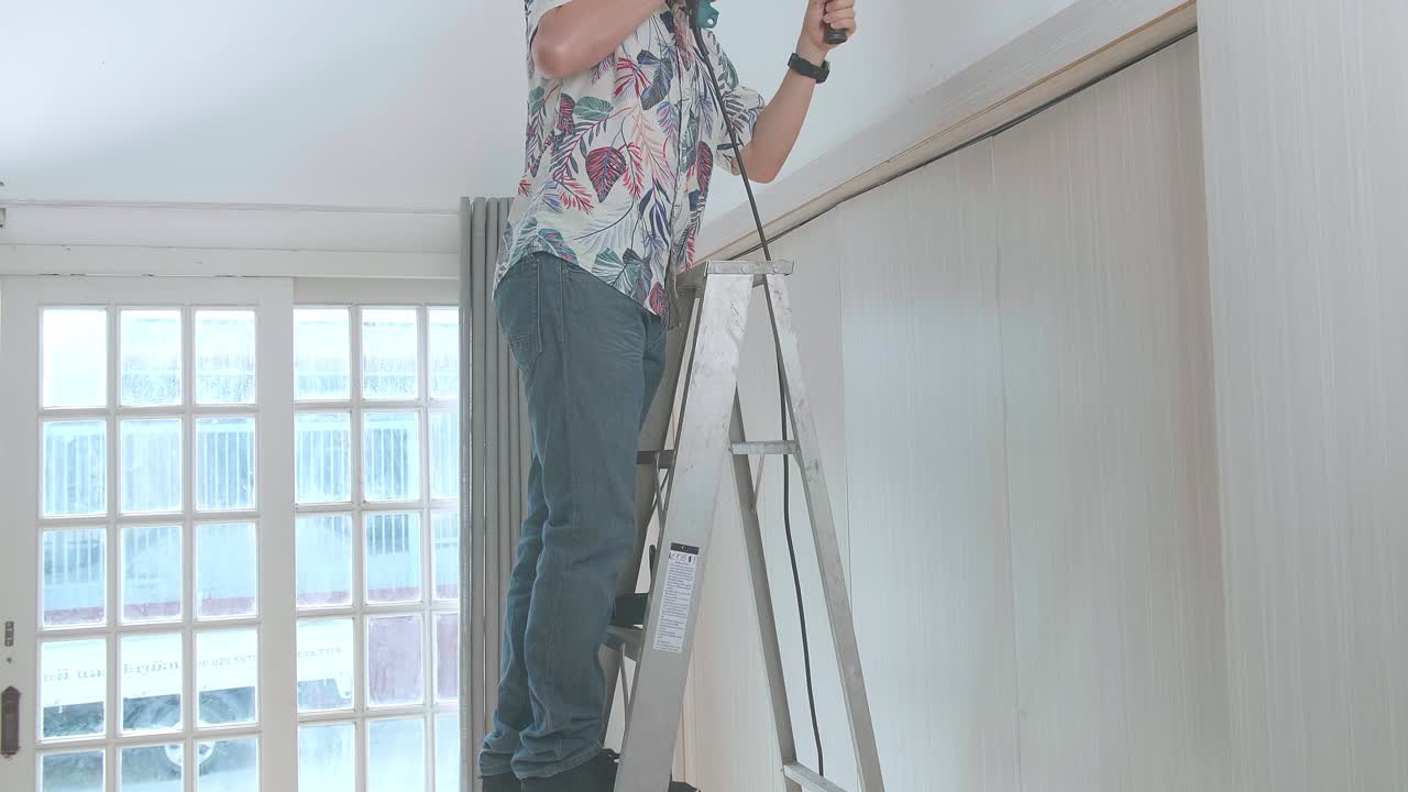 hombre asiático en una escalera usa taladro eléctrico en la pared