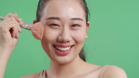 close up of beautiful asian woman rouging her cheeks while smiling to camera on green screen background in the studio