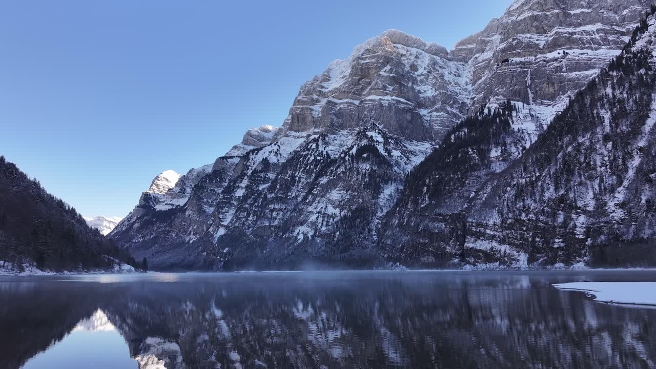 Klöntalersee in Switzerland. The crystal-clear lake reflects the snow-capped mountains that rise dramatically on either side, creating a mesmerizing mirror image.