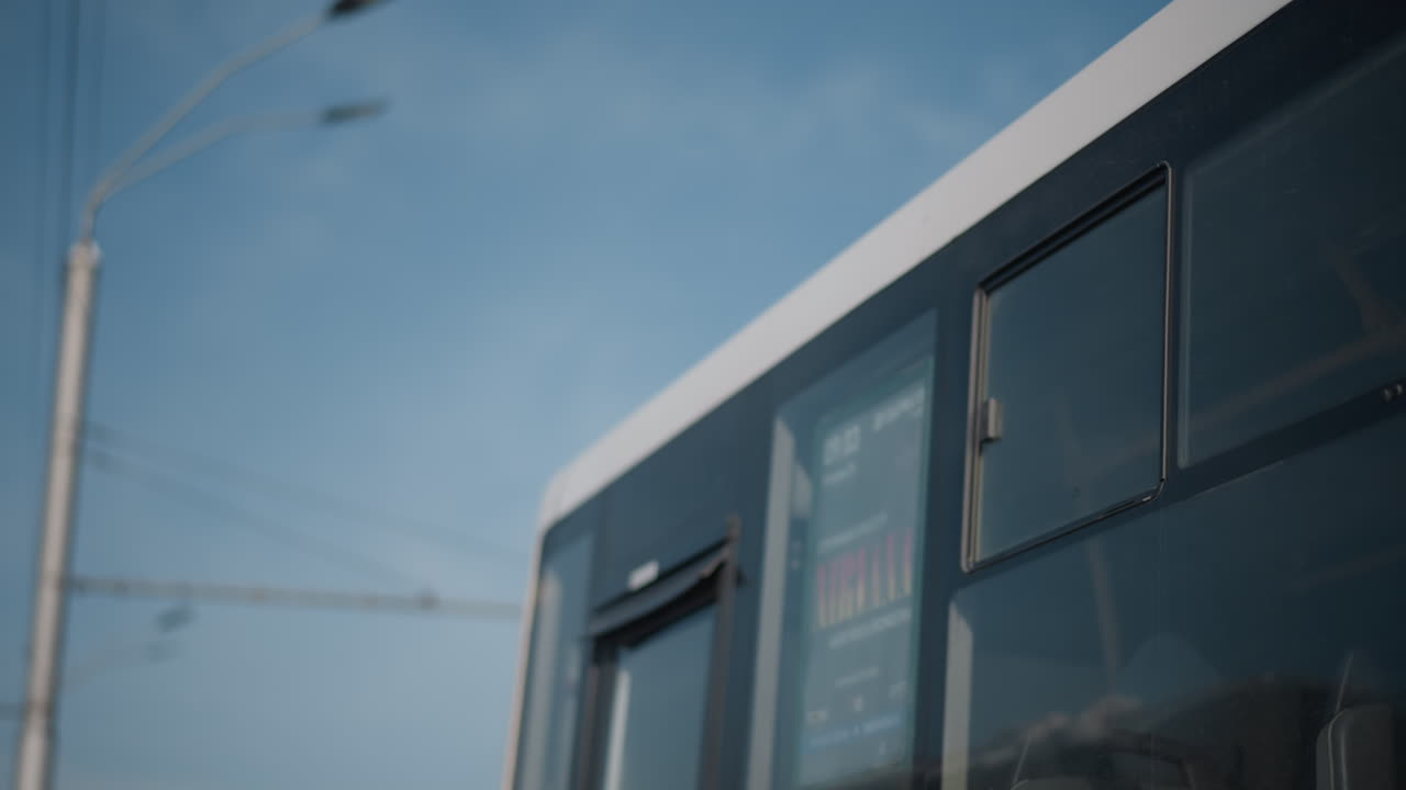 upward perspective of long city bus at stop, doors open for passengers, then close as vehicle departs, mirror and windows gleam under blue sky, wires and lamps frame transit scene