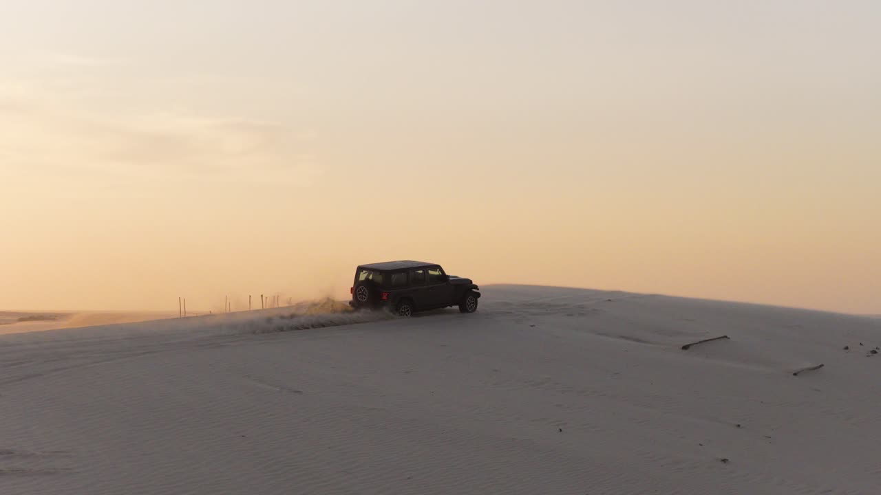 Una ripresa professionale in panning e tracking segue un veicolo 4x4 scuro mentre supera la cresta di una duna di sabbia bianca a Guriú, Brasile. Il tramonto splendente crea un bagliore dorato attraverso l'ampio paesaggio desertico.