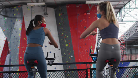 Two-diverse-women-wearing-face-masks-touching-elbows-at-gym