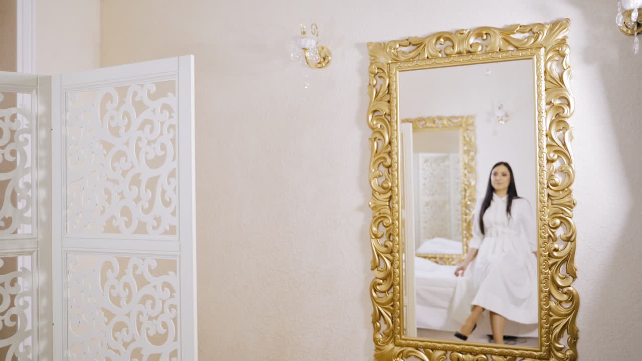 An elegant woman in a good mood with long black hair and a white dress came into the room to a specialist in permanent makeup and sat on the couch opposite the rectangular mirror in anticipation of the master.