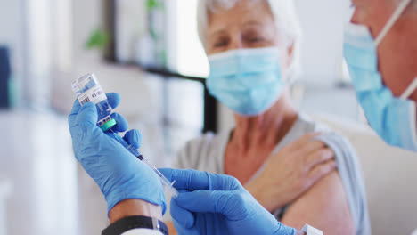 male caucasian doctor filling syringe with medication while senior caucasian woman wearing face mask