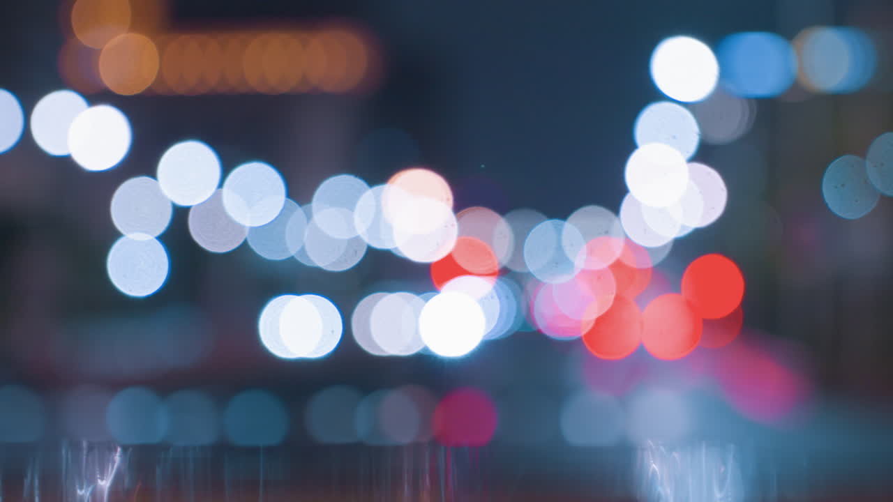 Luci bokeh sfocate della città su pavimentazione bagnata, fari e fanali posteriori fuori fuoco formano morbidi cerchi luminosi, toni blu freddi e arancioni caldi, riflessi scintillanti, texture cinematografica sognante ideale.