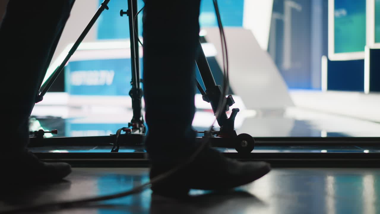 Close-up of studio camera tripod with operator s feet visible, camera dolly on track, lighting cables, set background blurred, broadcasting equipment in modern TV studio