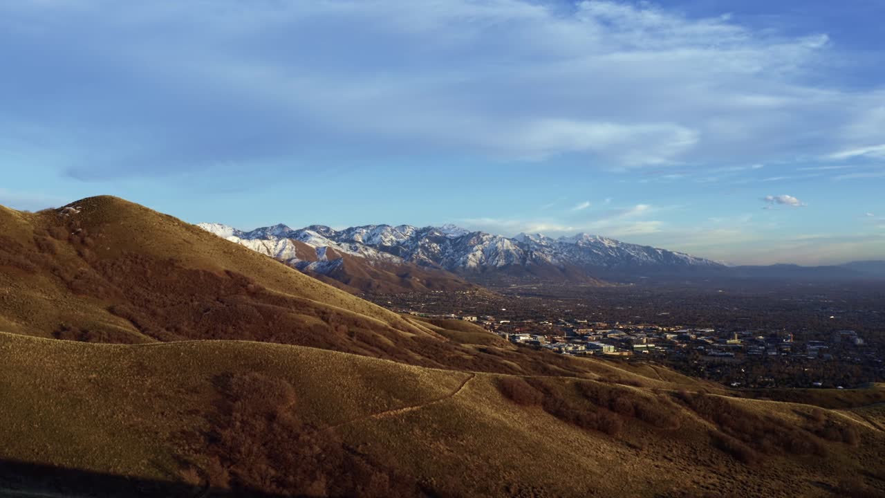 Wide aerial drone shot dollying out over the dry rolling hills of the Bonneville Shoreline trail in Salt Lake City, Utah looking out at the valley surrounded by large snow capped rocky mountains
