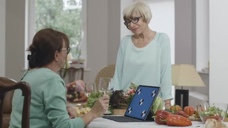 two positive senior women talking, cooking and surfing internet at home indoors. smiling beautiful caucasian retirees meeting indoors. laptop with blue screen on table. cinema 4k prores hq.