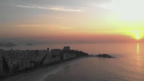 Paneo-Aéreo-Que-Muestra-El-Sol-Saliendo-Sobre-El-Horizonte-Del-Océano-Detrás-De-Arpoador-Revelando-La-Playa-De-Ipanema-Vacía-Temprano-En-La-Mañana-Más-Ancha-En-Primer-Plano-Y-El-Paisaje-Urbano-Más-Amplio-De-Río-De-Janeiro-En-La-Parte-Posterior